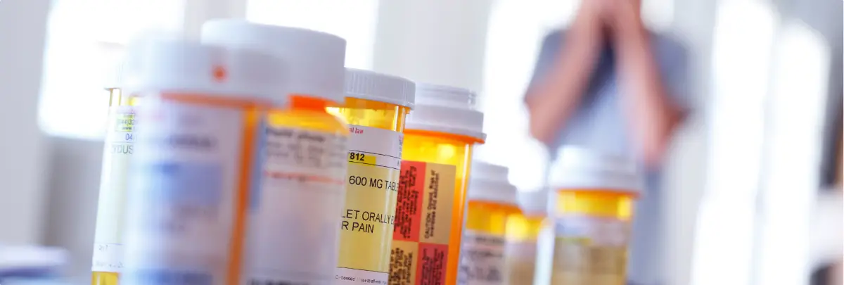 Row of prescription pill bottles in focus with a distressed person blurred in the background.